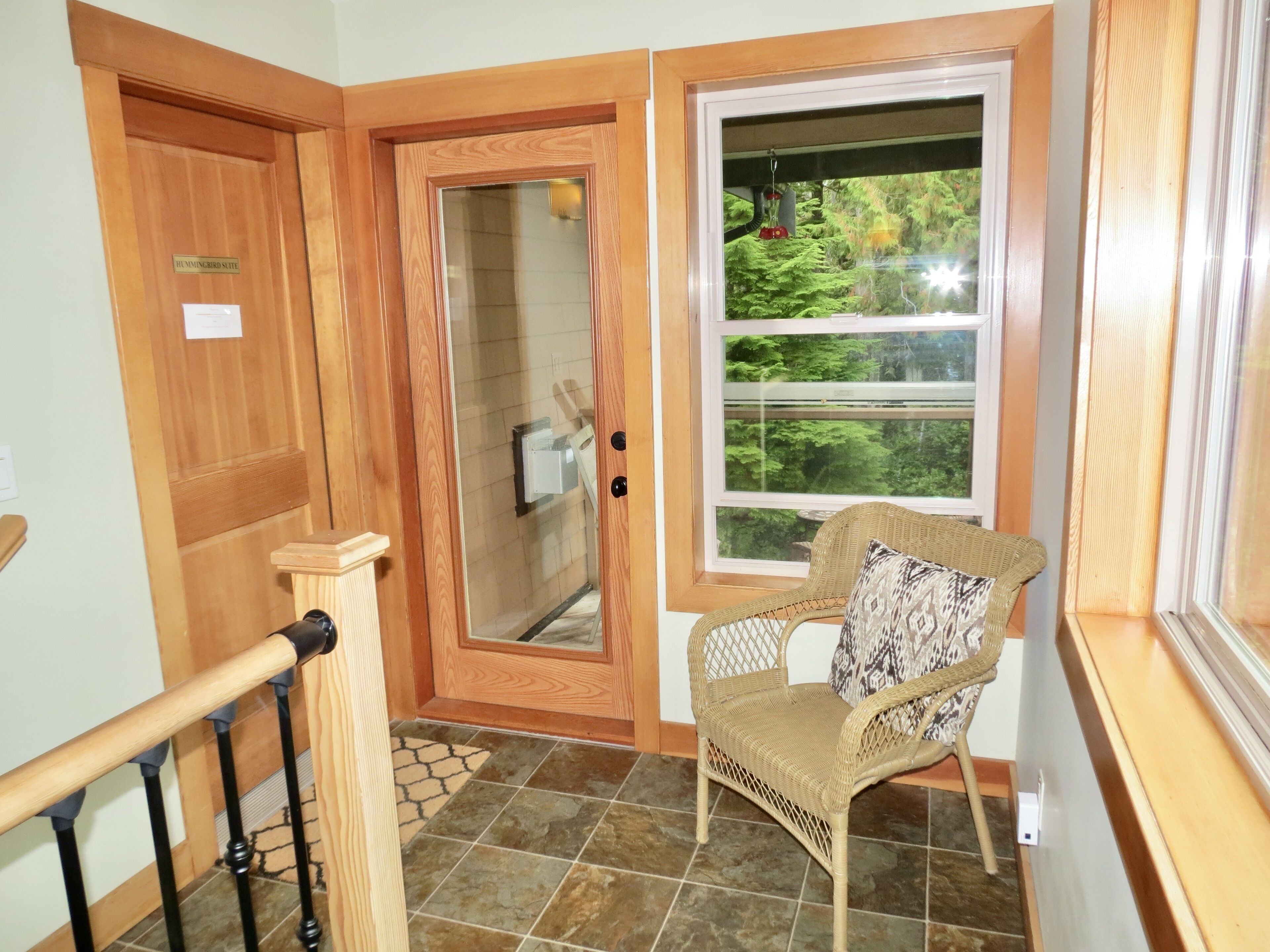 A wooden door labeled "Hummingbird Suite," a wicker chair with a patterned cushion, and a window with a view of greenery.