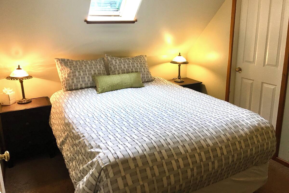 Um quarto com uma cama, duas mesas de cabeceira com abajures, uma janela e uma porta.
