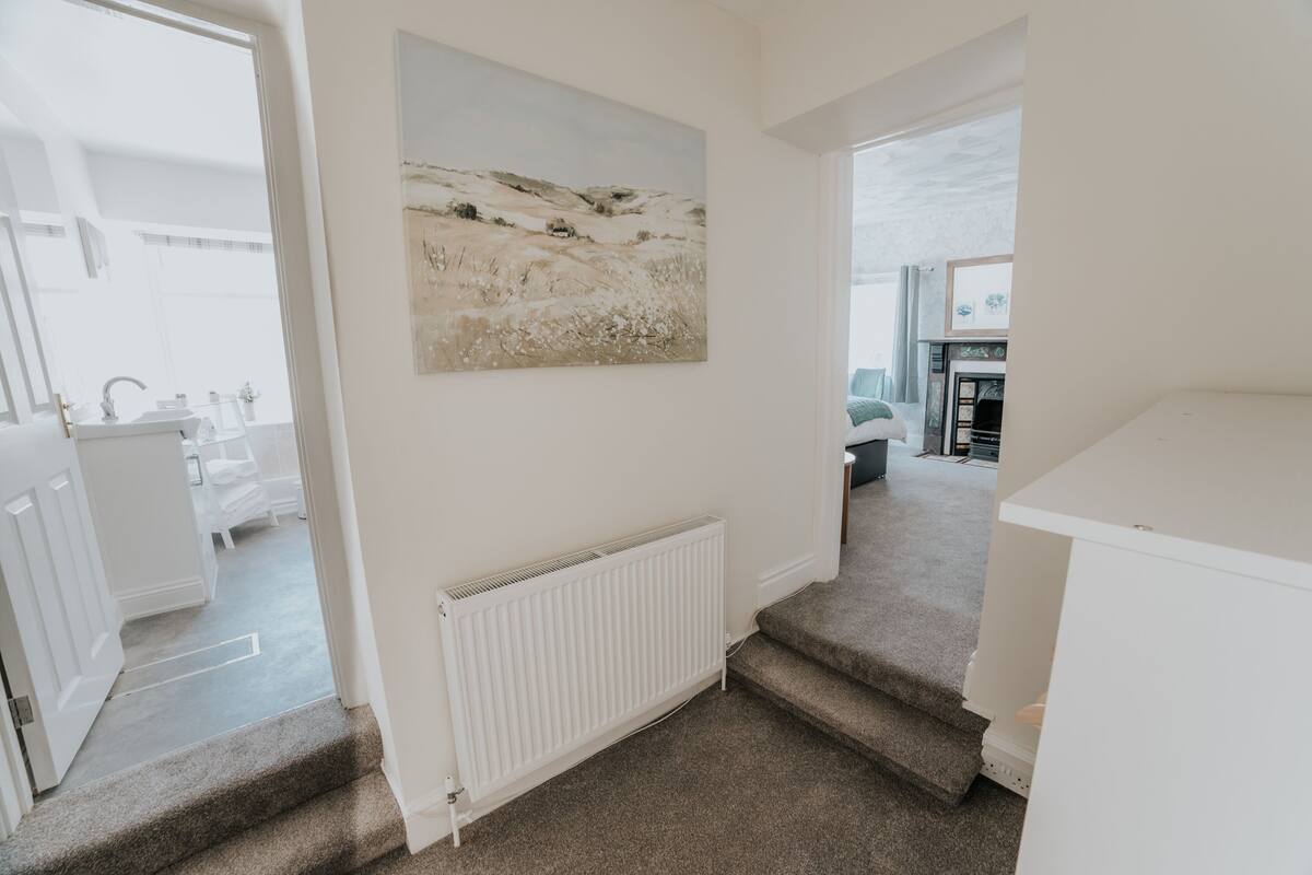 Un couloir avec un escalier recouvert de moquette, un radiateur blanc et un tableau représentant un paysage accroché au mur.