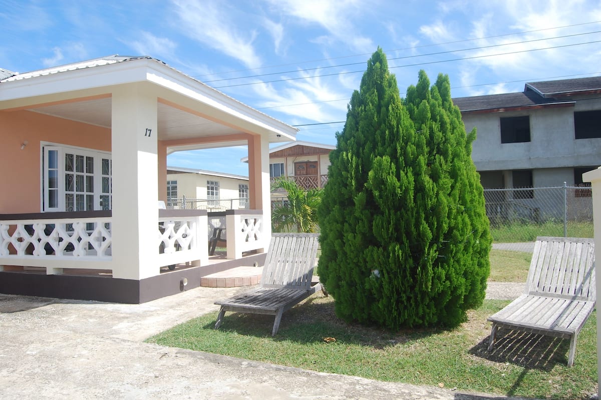 A covered outdoor area with two wooden benches, a small tree, and a building with a balcony.
