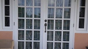 A covered patio with a white door, two windows with decorative grills, and a welcome mat. - Sue'S Homestay (Oistins)