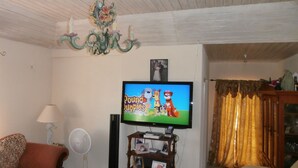 A living room with a television displaying 'Pound Puppies', a chandelier, a fan, and a wooden cabinet with photos. - Sue'S Homestay (Oistins)