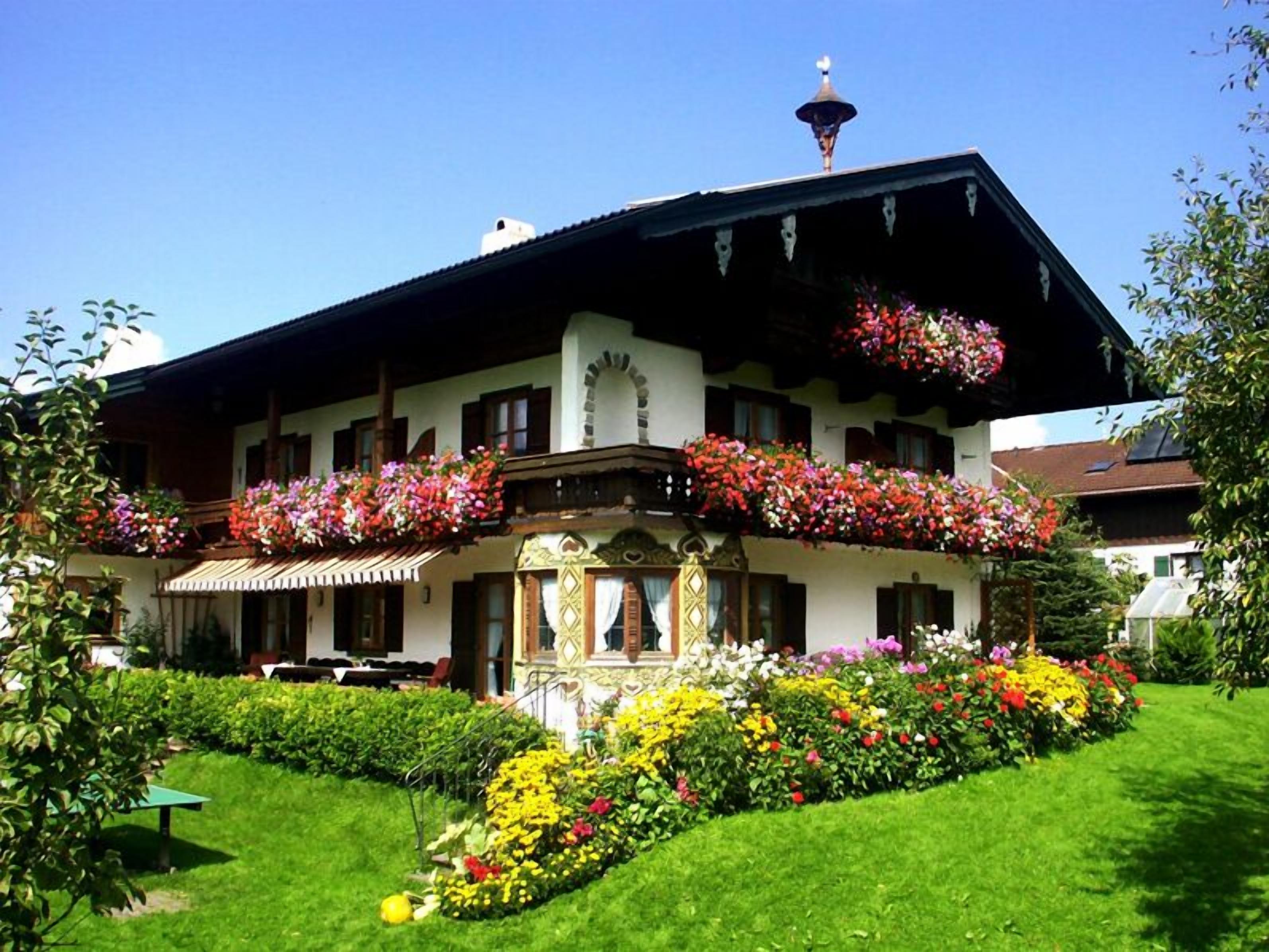 A traditional Alpine-style building with a balcony adorned with colorful flowers, a well-maintained garden, and a clear blue sky.