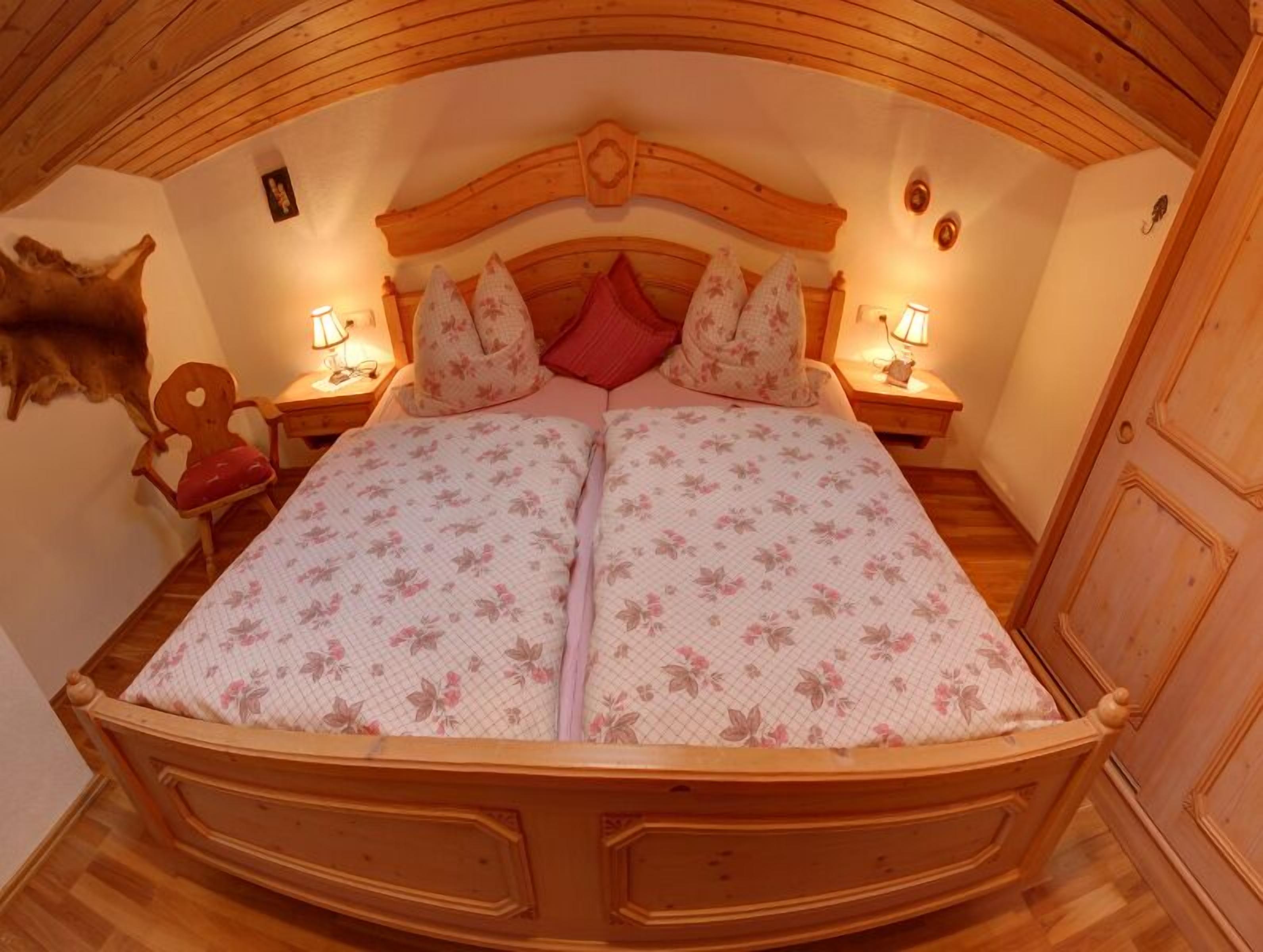 Een knus vertrek met een houten plafond, een bed met bloemenbedrukking, twee nachtkastjes met lampen, en een stoel.
