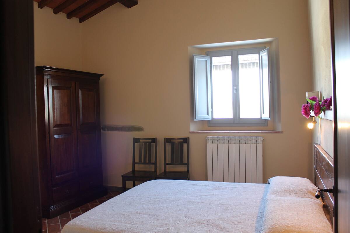 Un dormitorio con cama, dos sillas, un armario de madera, una ventana y un radiador.