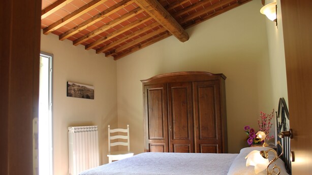 Une chambre avec un plafond en bois, un lit recouvert d’une courtepointe blanche, une armoire en bois, une chaise blanche et une table de chevet avec une lampe.