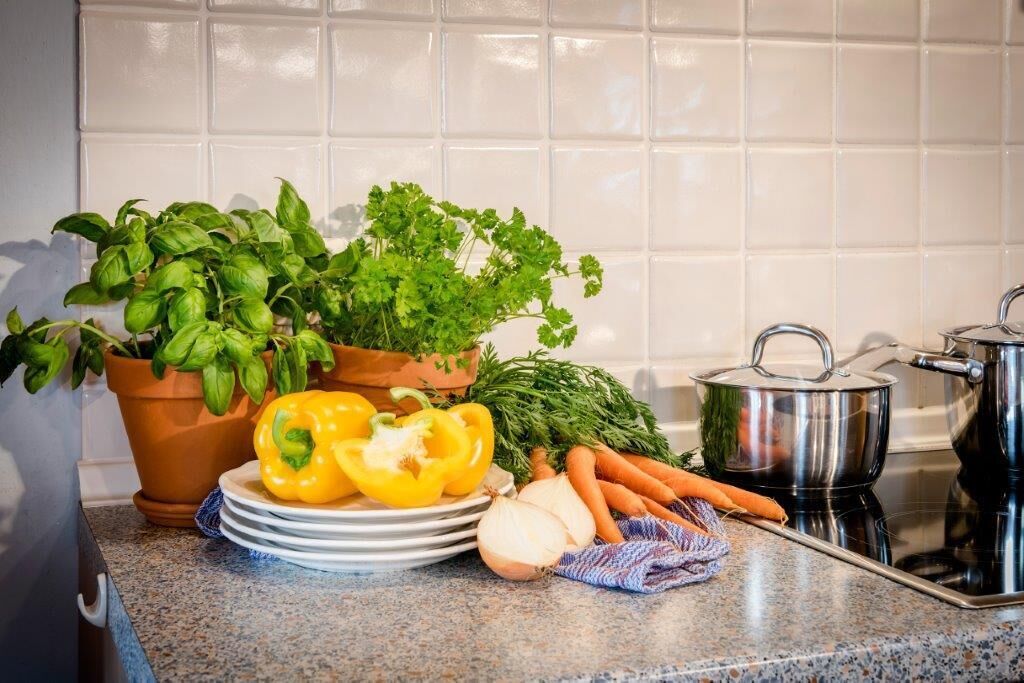 A kitchen counter with fresh vegetables, a potted basil plant, a yellow bell pepper, a carrot, a plate, a pot, and a stove.