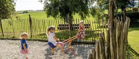 Niños jugando en un sube y baja en un área exterior delimitada, con un campo de césped y árboles al fondo.