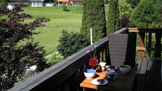 Une terrasse en bois avec une table dressée pour le repas, donnant sur un champ verdoyant et sur des maisons en arrière-plan.