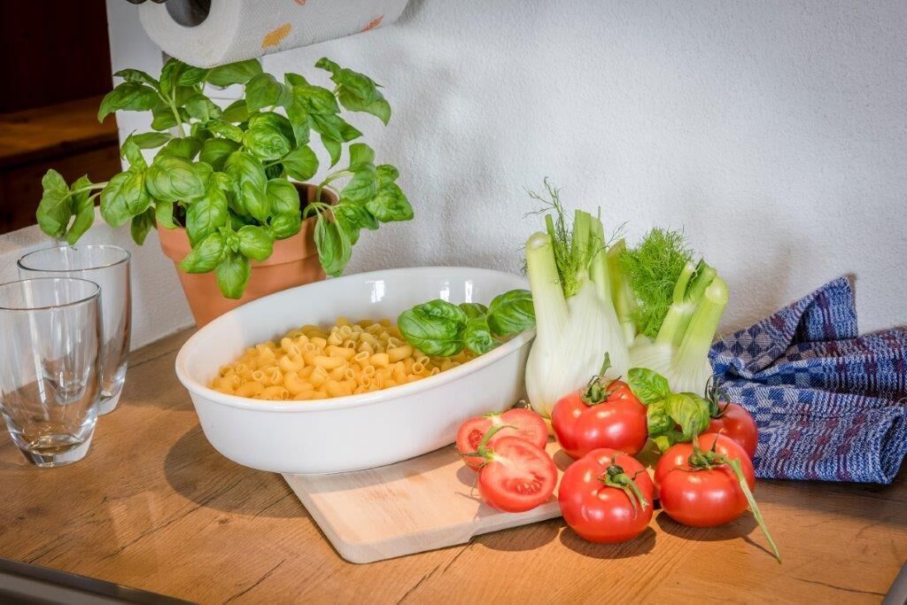 A kitchen counter with a bowl of pasta, fresh vegetables, and a potted basil plant.