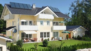 Una casa de dos pisos con un balcón, un patio con mobiliario exterior y paneles solares en el techo.