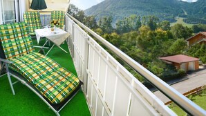 Un balcón con un sofá verde a cuadros, una mesa pequeña y un parasol verde. El balcón ofrece vistas a un paisaje montañoso con árboles y un edificio.