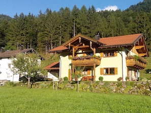 A two-story chalet with wooden balconies, a stone foundation, and a mountainous backdrop.