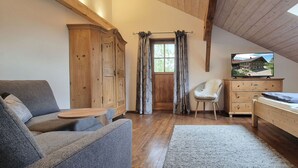 A cozy room with a wooden ceiling, a sofa, a chair, a bed, a TV, and a window with curtains. - Ferienwohnung Bauernhaus Hochgernblick (Grassau)