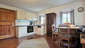 A compact kitchen with wooden cabinets, a dining area with a bench, and a window with curtains. - Ferienwohnung Bauernhaus Hochgernblick (Grassau)