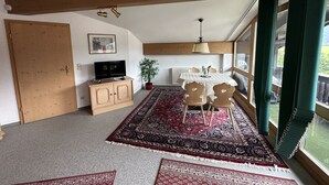 Una habitación con techo de madera, un televisor sobre un mueble, una mesa de comedor con sillas, una alfombra estampada y una ventana con cortinas.