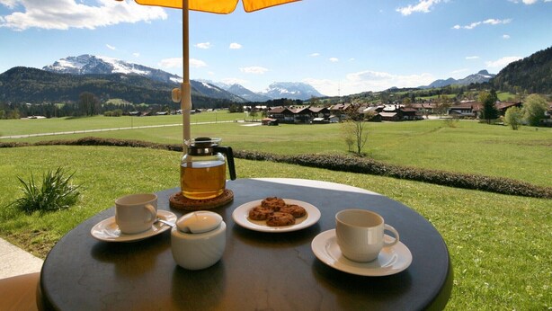 Ett bord med en kaffekanna, koppar och kakor under ett gult parasoll, med utsikt över en pittoresk by med berg i bakgrunden.