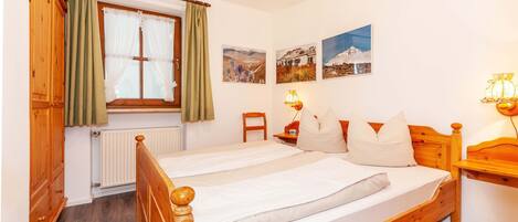 A bedroom with a wooden bed, a radiator, a window with curtains, a chair, and wall-mounted lamps.
