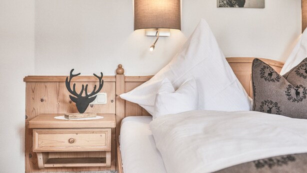Un lit bien fait, avec une literie blanche, une table de chevet en bois ornée d’une tête de cerf, une lampe fixée au mur et un tableau représentant un cerf, encadré.