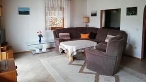 Una sala acogedora con un sofá marrón, una mesa de centro de vidrio y un mantel blanco. Se ve una ventana con cortinas blancas, una lámpara y cuadros enmarcados en la pared.