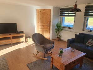 Uma sala de estar com um móvel de entretenimento de madeira, uma poltrona cinza, uma cadeira de balanço, uma mesa de centro de madeira e um sofá cinza.