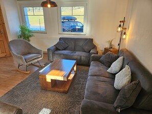 A living room with a grey sofa, a wooden coffee table, a rocking chair, and a view of a car through a window. - Komfortable Ferienwohnung, 85 qm, 2 sep. Schlafzimmer und Küche (Reit im Winkl)