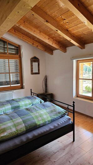 A bedroom with a wooden ceiling, a bed with green and gray bedding, a radiator, and a window with blinds. - Komfortable Ferienwohnung, 85 qm, 2 sep. Schlafzimmer und Küche (Reit im Winkl)