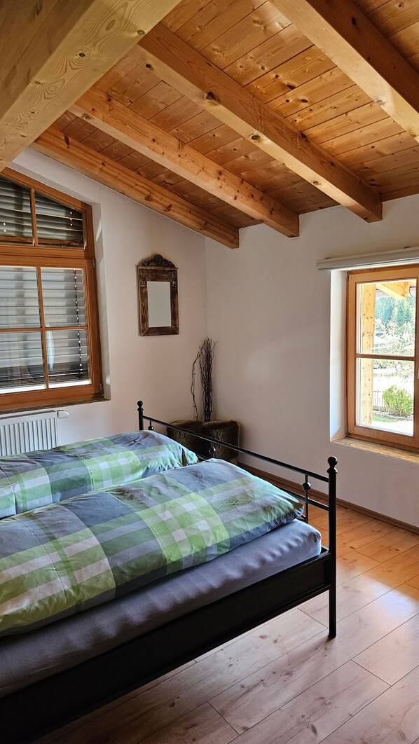 A bedroom with a wooden ceiling, a bed with green and gray bedding, a radiator, and a window with blinds.