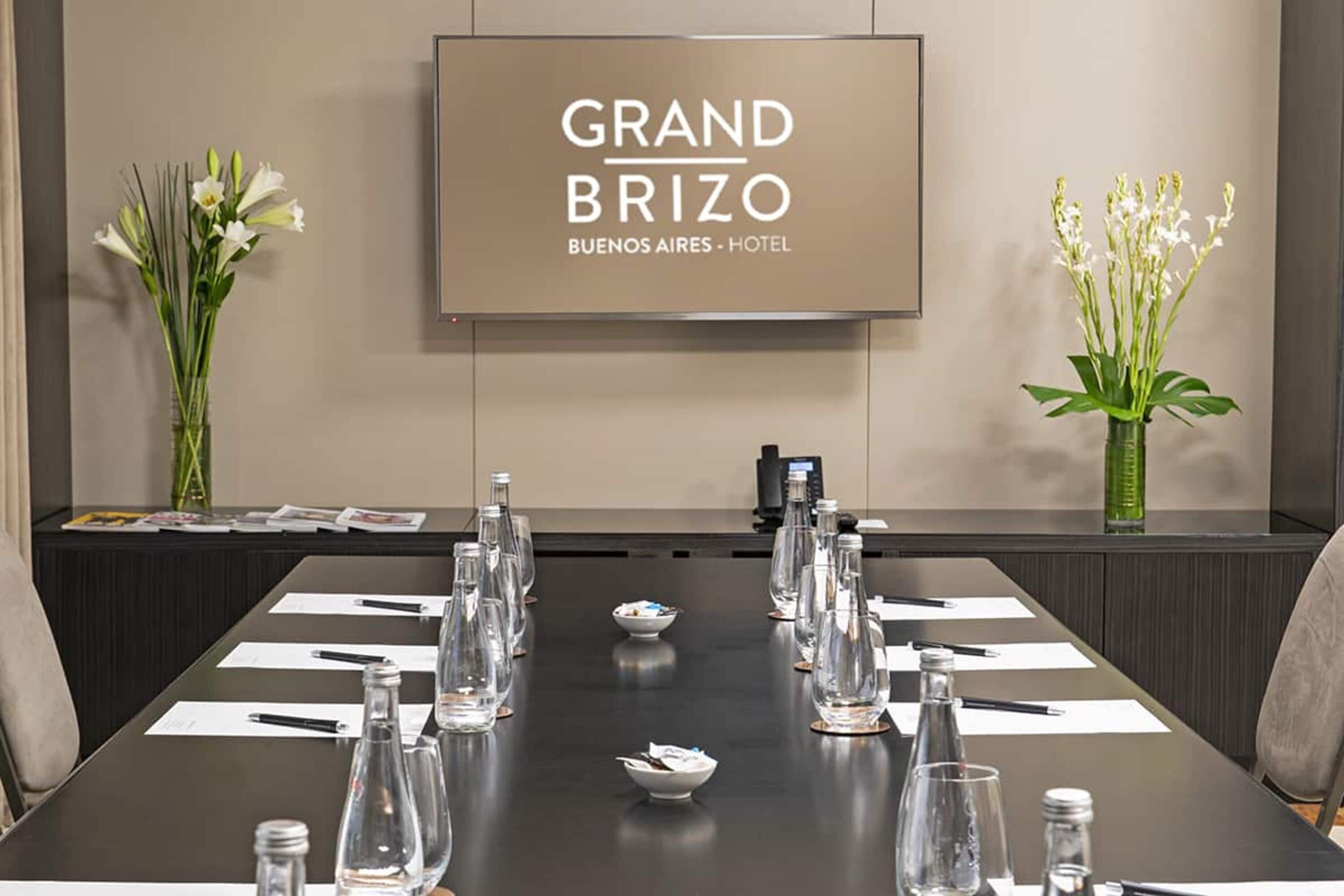 A conference room at the Grand Brizo Buenos Aires Hotel with a large screen displaying the hotel's name, a long table set with water bottles, glasses, and name cards.