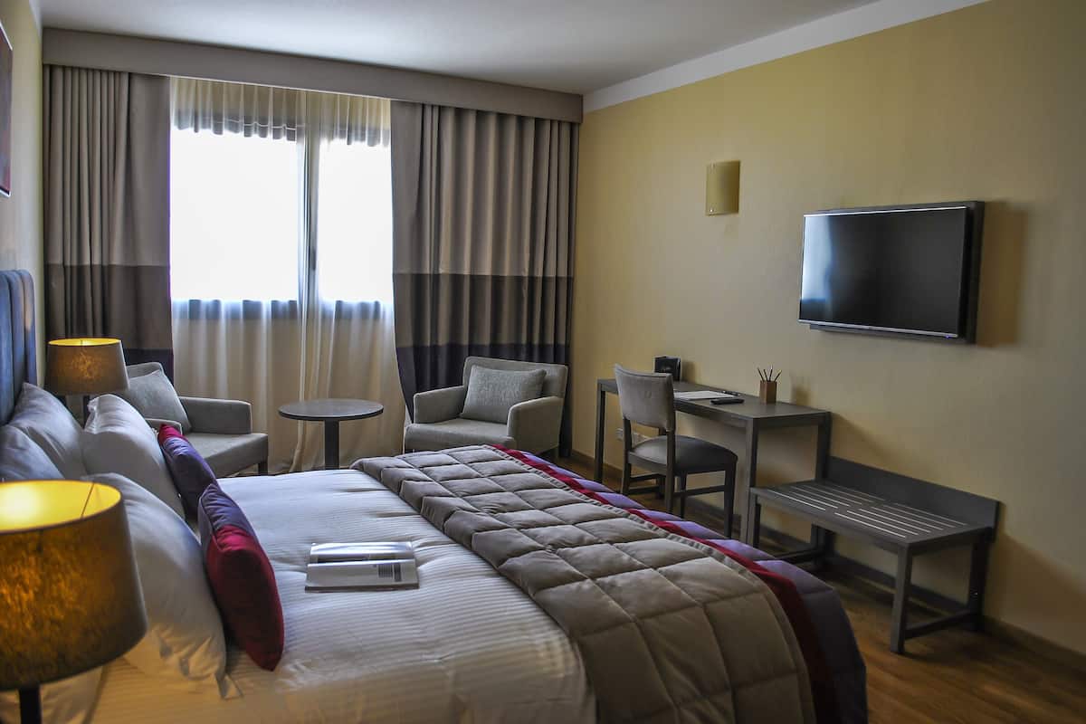 A hotel room with a bed, a desk, a chair, a TV, and a window with curtains.