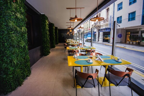 An outdoor dining area with yellow tables and chairs, a green wall, and a view of the street. - Grand Brizo Buenos Aires (Buenos Aires)