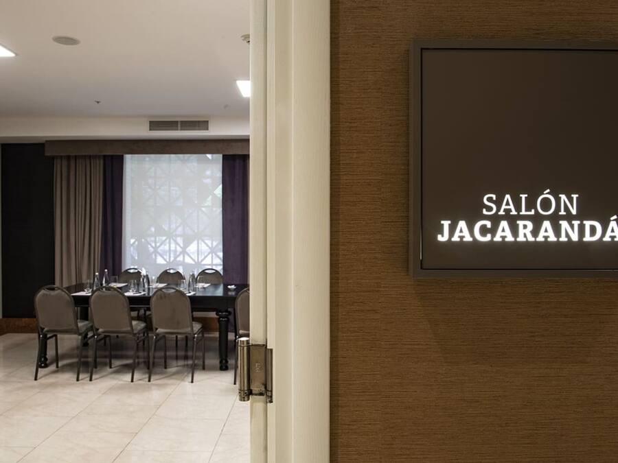 A conference room with a table and chairs, a sign reading "SALÓN JACARANDA," and a window with curtains.