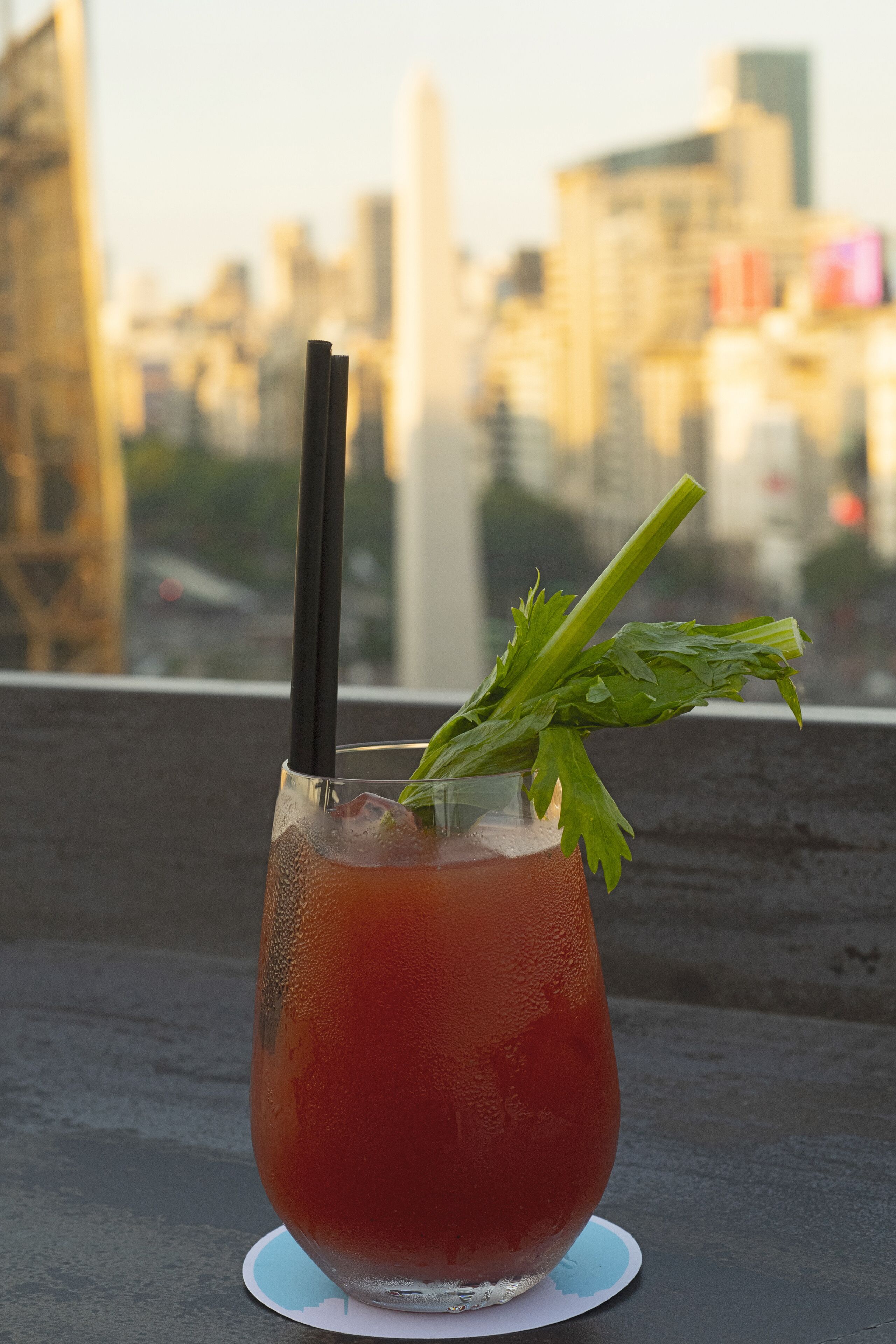 A glass of red beverage with a black straw and celery garnish on a coaster, set against a cityscape background.