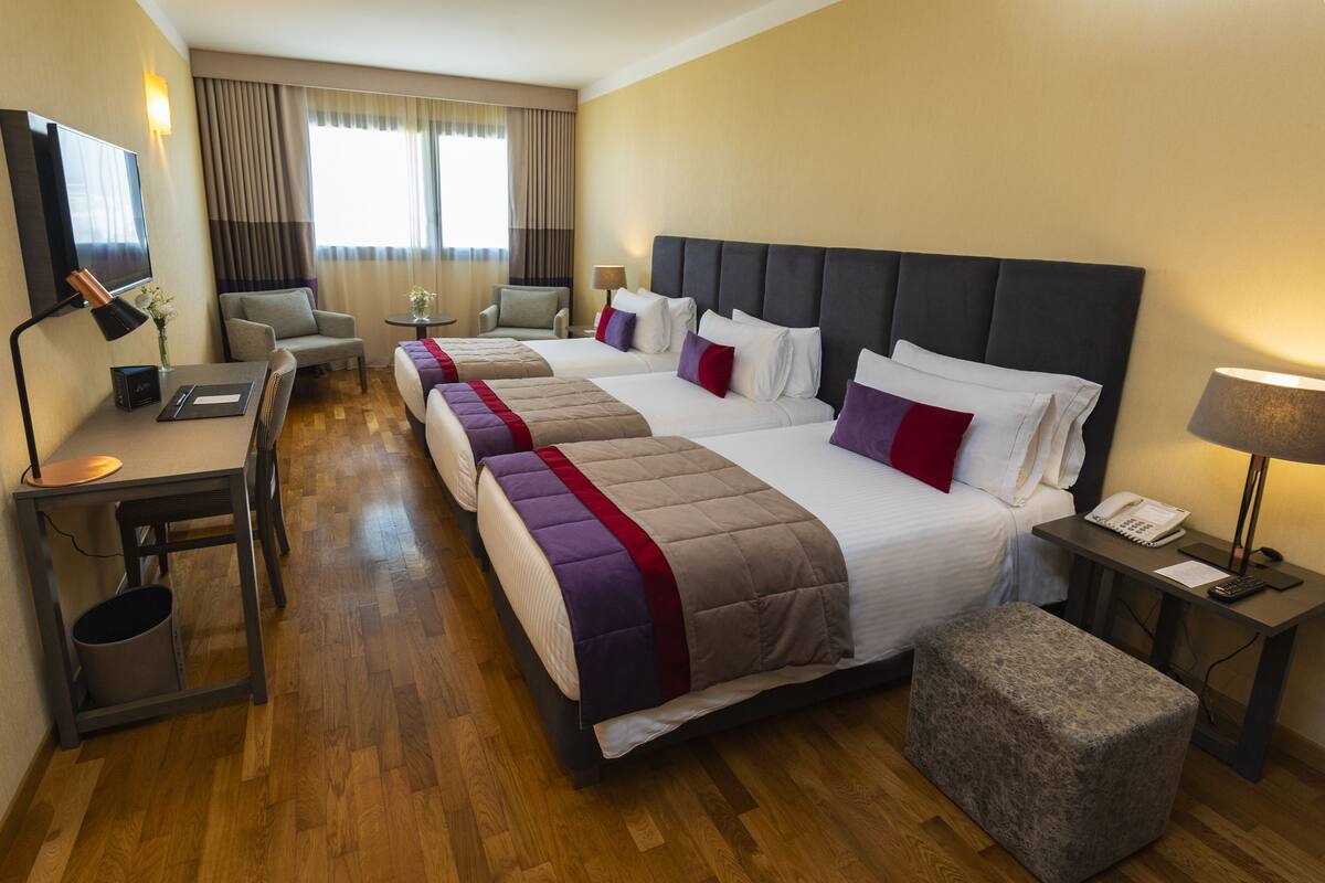 A hotel room with three beds, a desk, a chair, a TV, and a lamp.