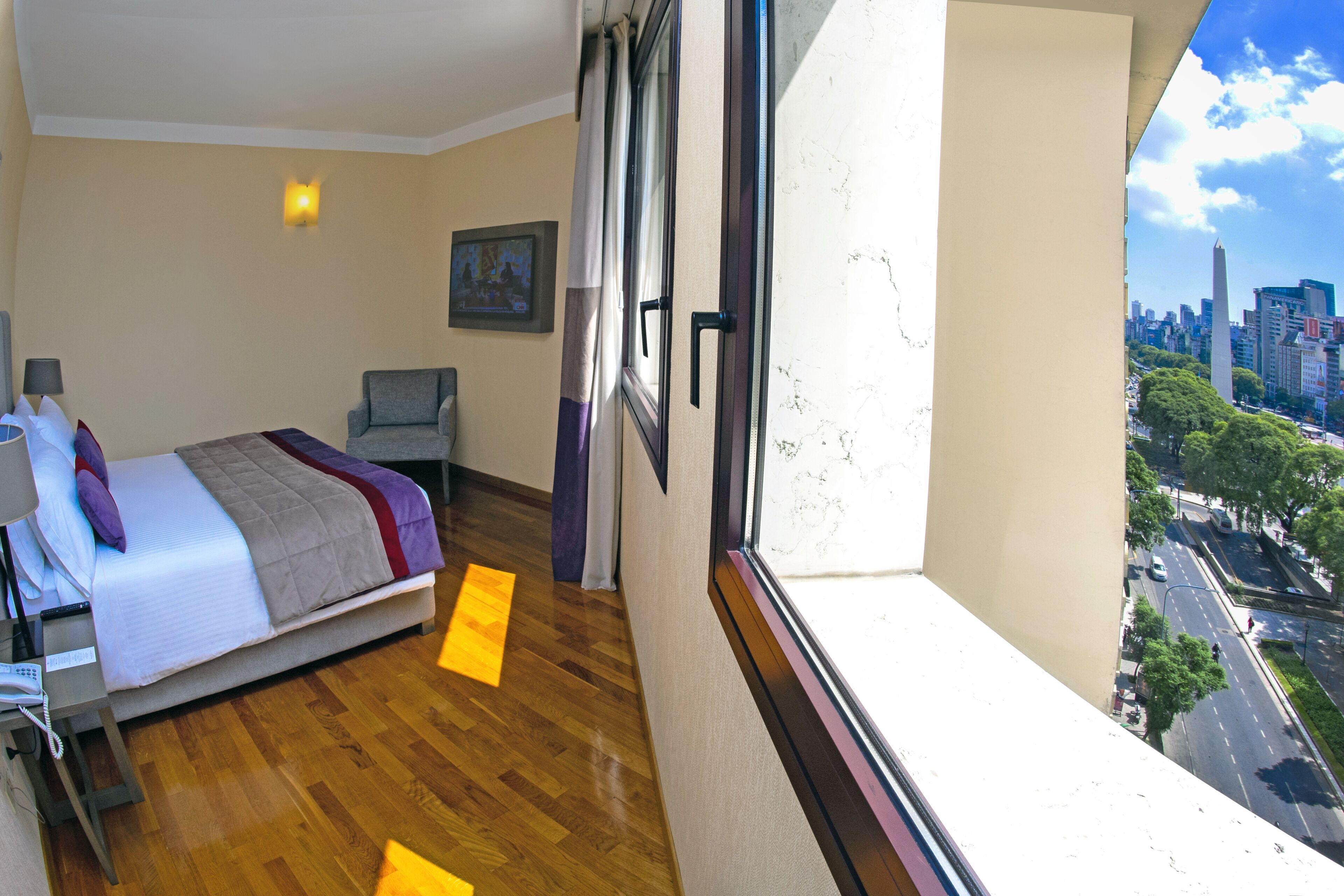 A hotel room with a bed, a desk with a lamp, a chair, and a window with a city view.