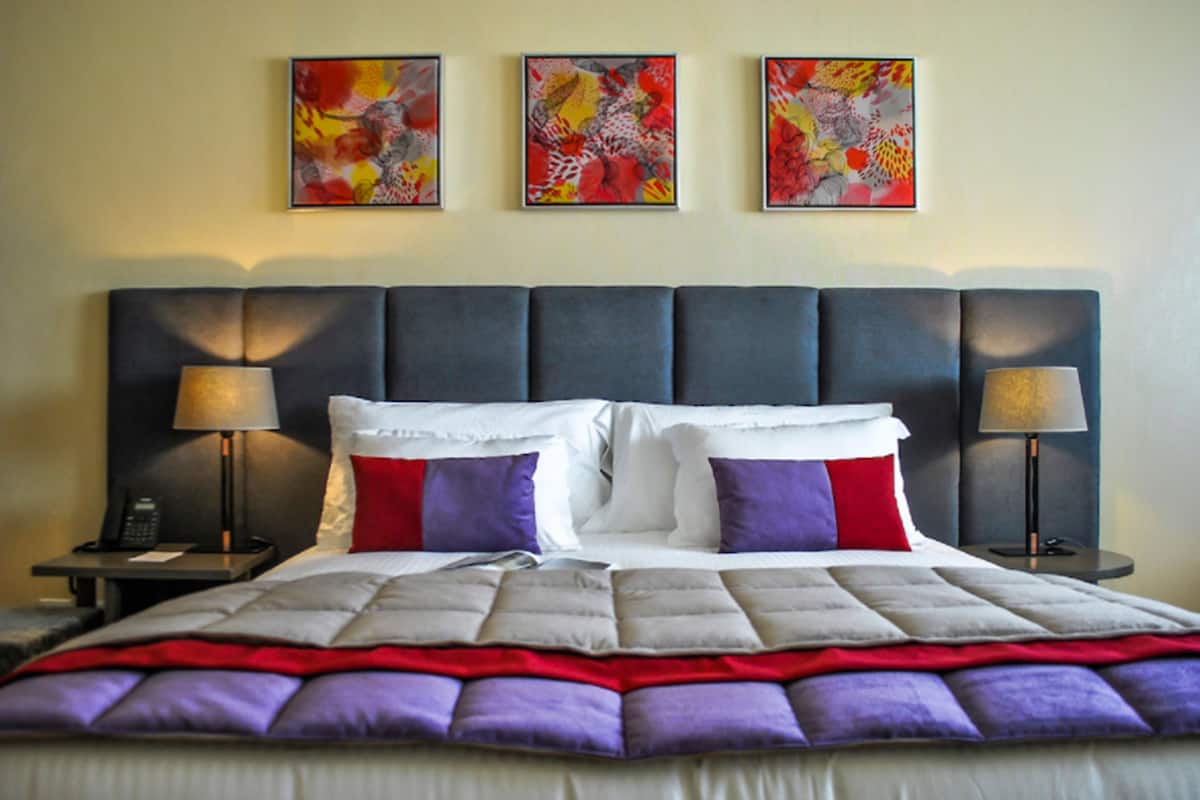 A hotel room with a large bed, two bedside tables with lamps, and three abstract paintings on the wall.