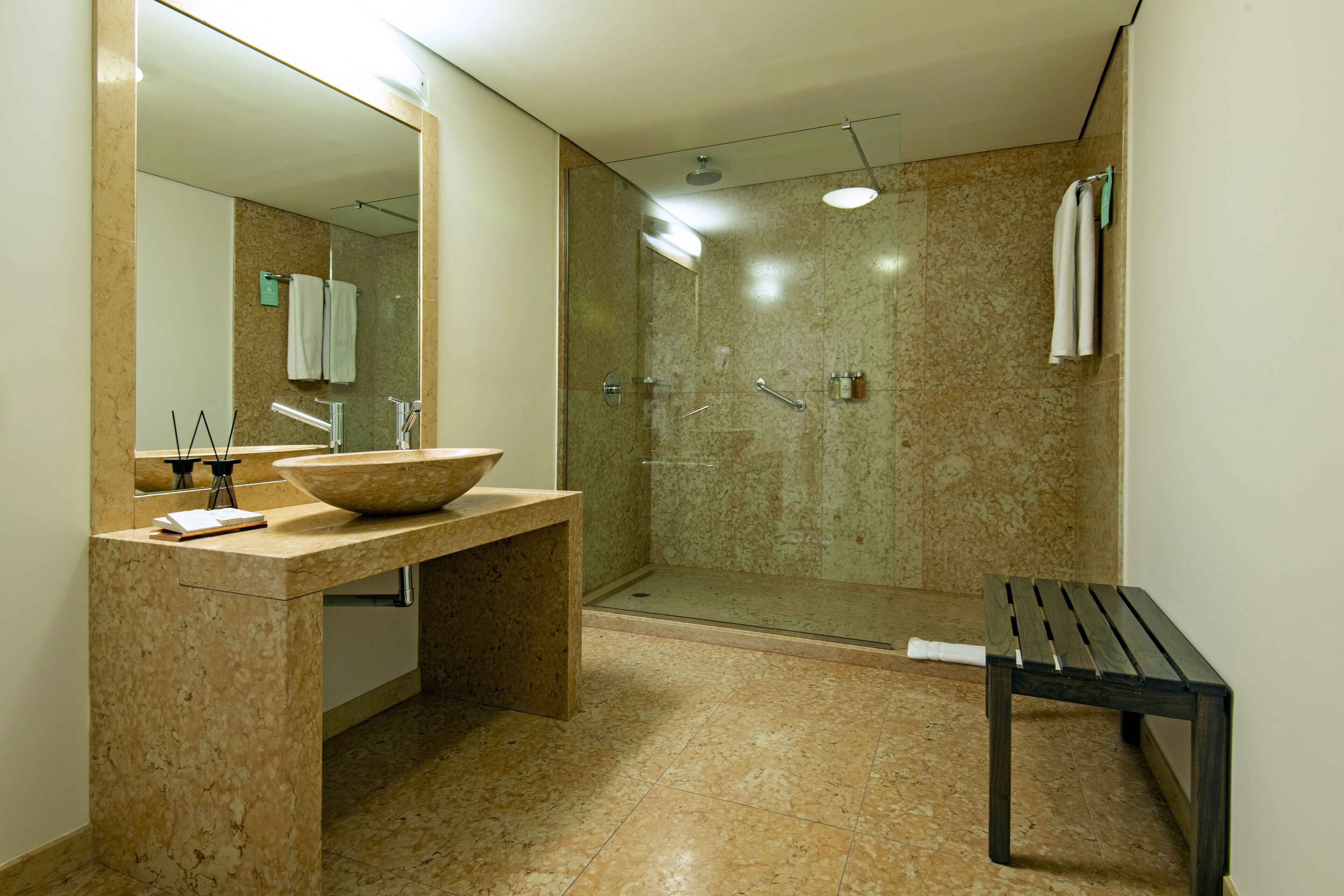 A bathroom with a large mirror, a sink with a bowl, a shower, and a bench.