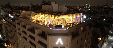 Un hotel con terraza en la azotea, diseño moderno, varios niveles y un llamativo letrero iluminado con la letra 'A' en el edificio.