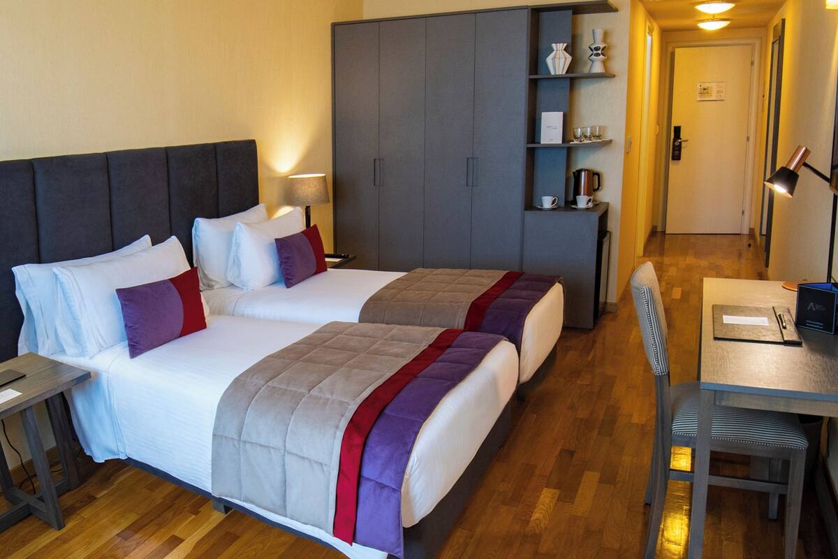 A hotel room with two beds, a desk, a chair, a wardrobe, and a shelf with items.