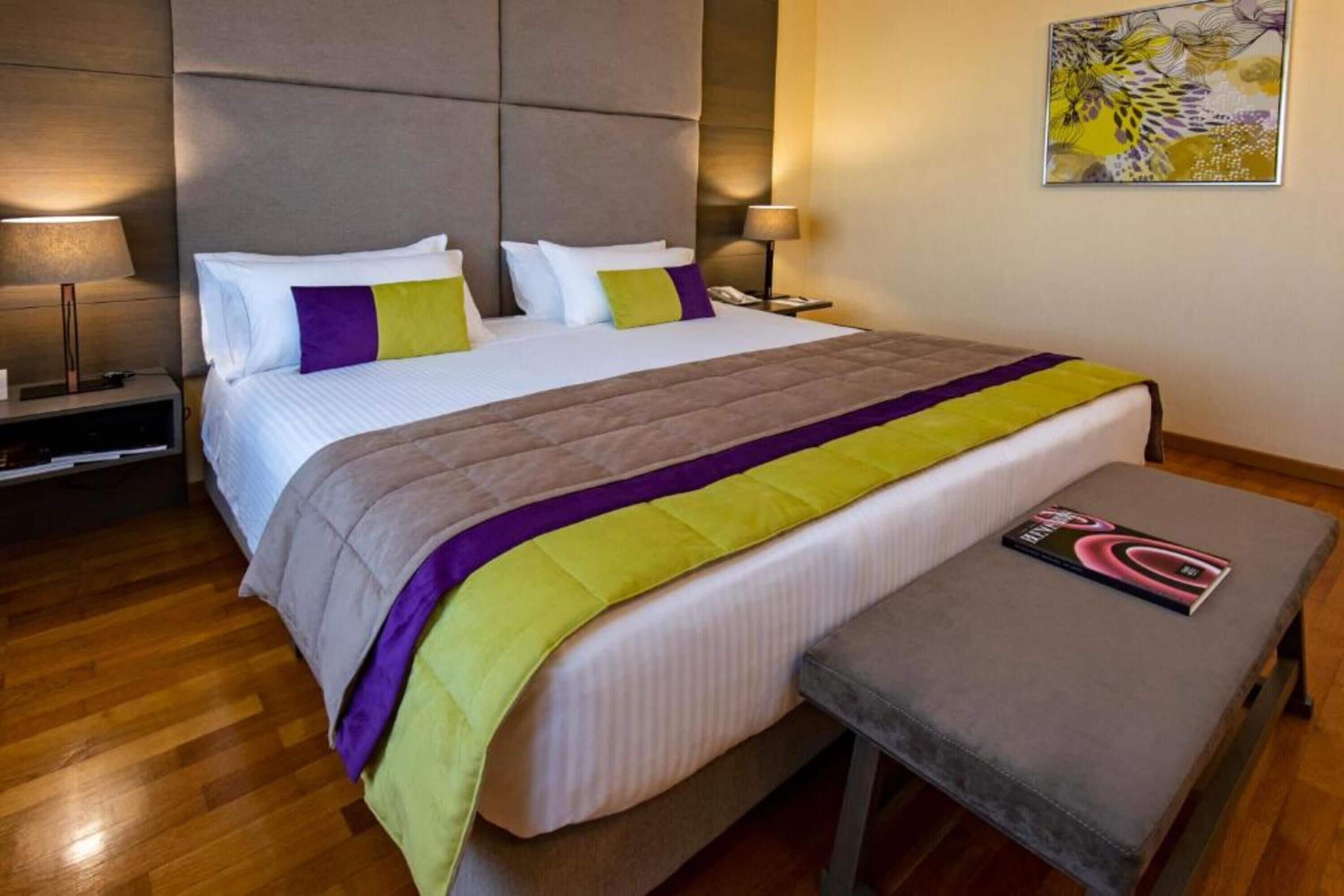 Una habitación de hotel con cama, mesitas de noche, un banco y una lámpara en la pared.