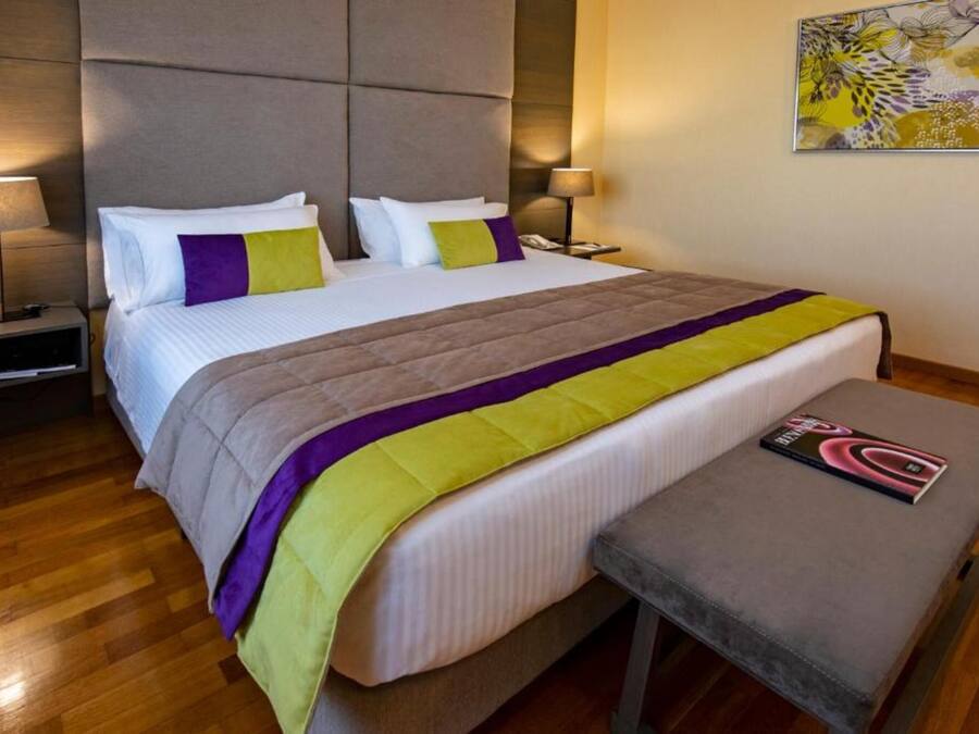 Una habitación de hotel con cama, mesitas de noche, un banco y una lámpara en la pared.