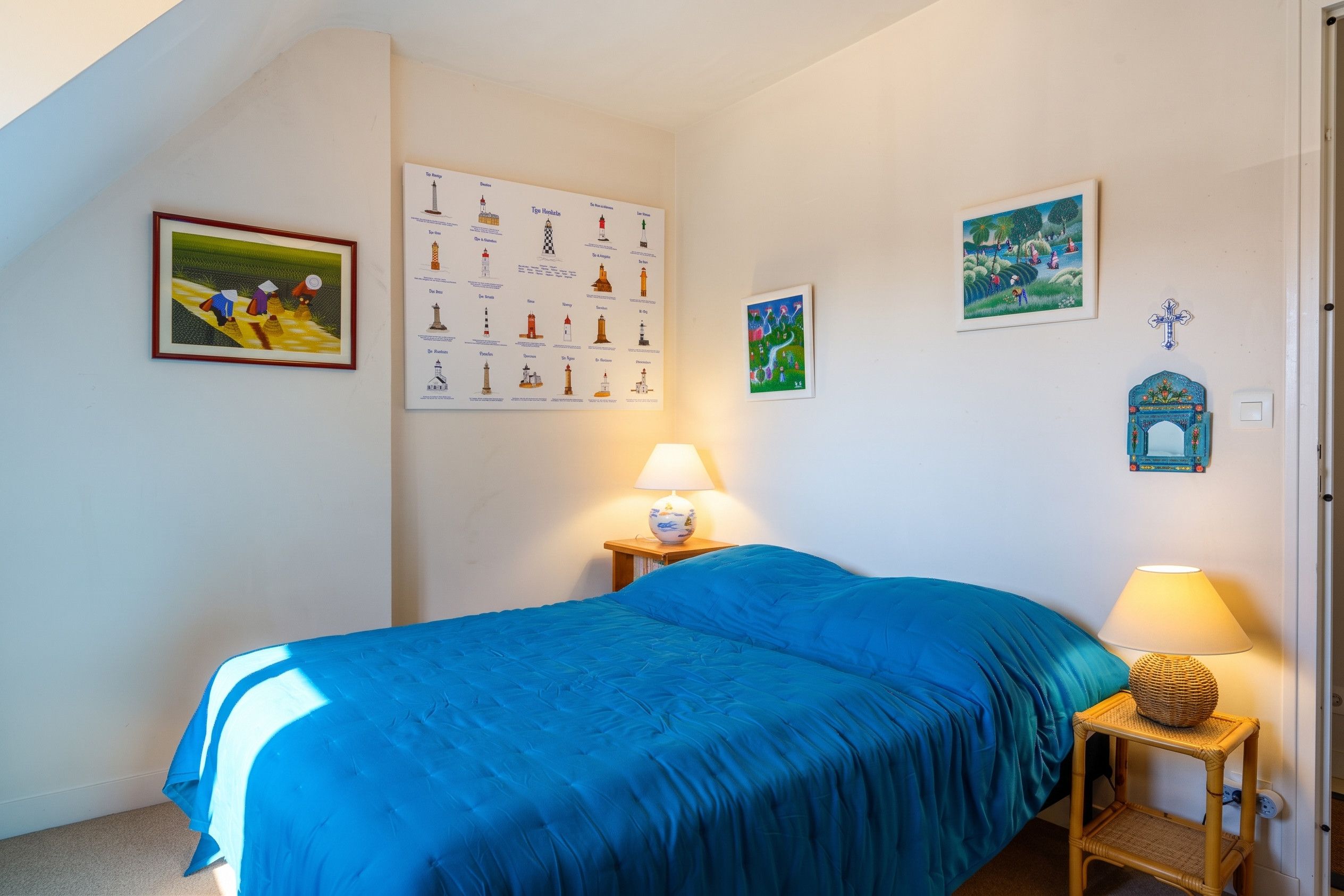 A bedroom with a bed, a nightstand, a lamp, and wall decorations including a lighthouse poster and a painting.