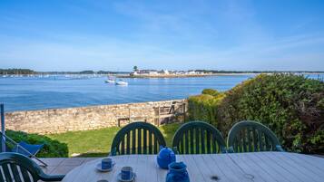Une table entourée de chaises, avec vue sur une mer calme où voguent des bateaux, un mur de pierre et une ville lointaine.