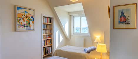 A bedroom with two beds, a bookshelf, a window with a view, and a hanging light fixture.