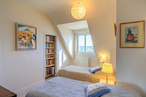 A bedroom with two beds, a bookshelf, a window with a view, and a hanging light fixture. - Homerez - “House in Loctudy, garden view.” (Loctudy)