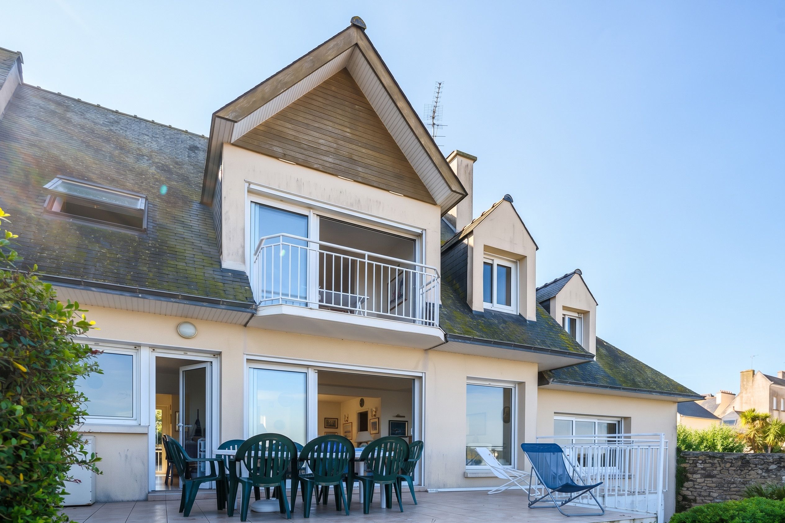 A two-story house with a balcony, a dining area with chairs, and a small outdoor seating area.