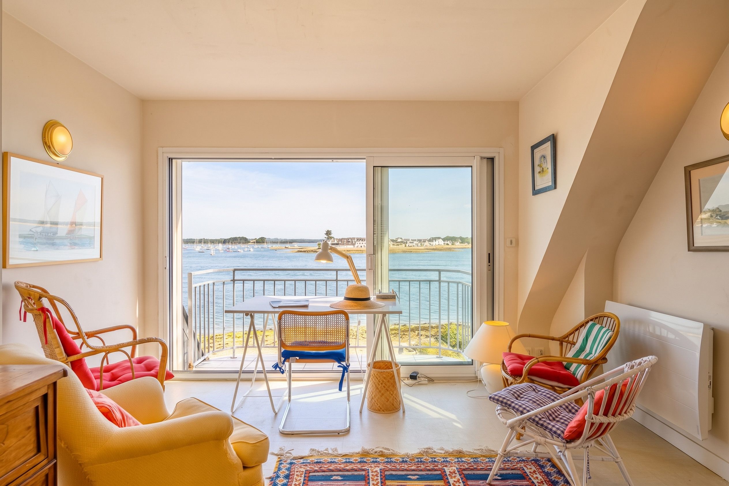 Ein Balkon mit Meerblick, Korbstühle, ein Tisch mit Lampe und ein gerahmtes Bild an der Wand.