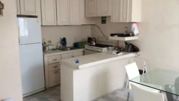 Una cocina con armarios blancos, un refrigerador, un lavamanos y una mesa de vidrio con sillas.