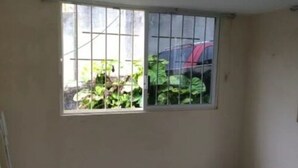 Una habitación con ventanal de barrotes, pared lisa y un coche rojo visible afuera.