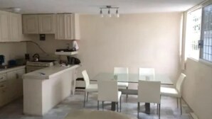 Una cocina compacta con armarios blancos y un pequeño comedor con mesa de vidrio y sillas blancas.